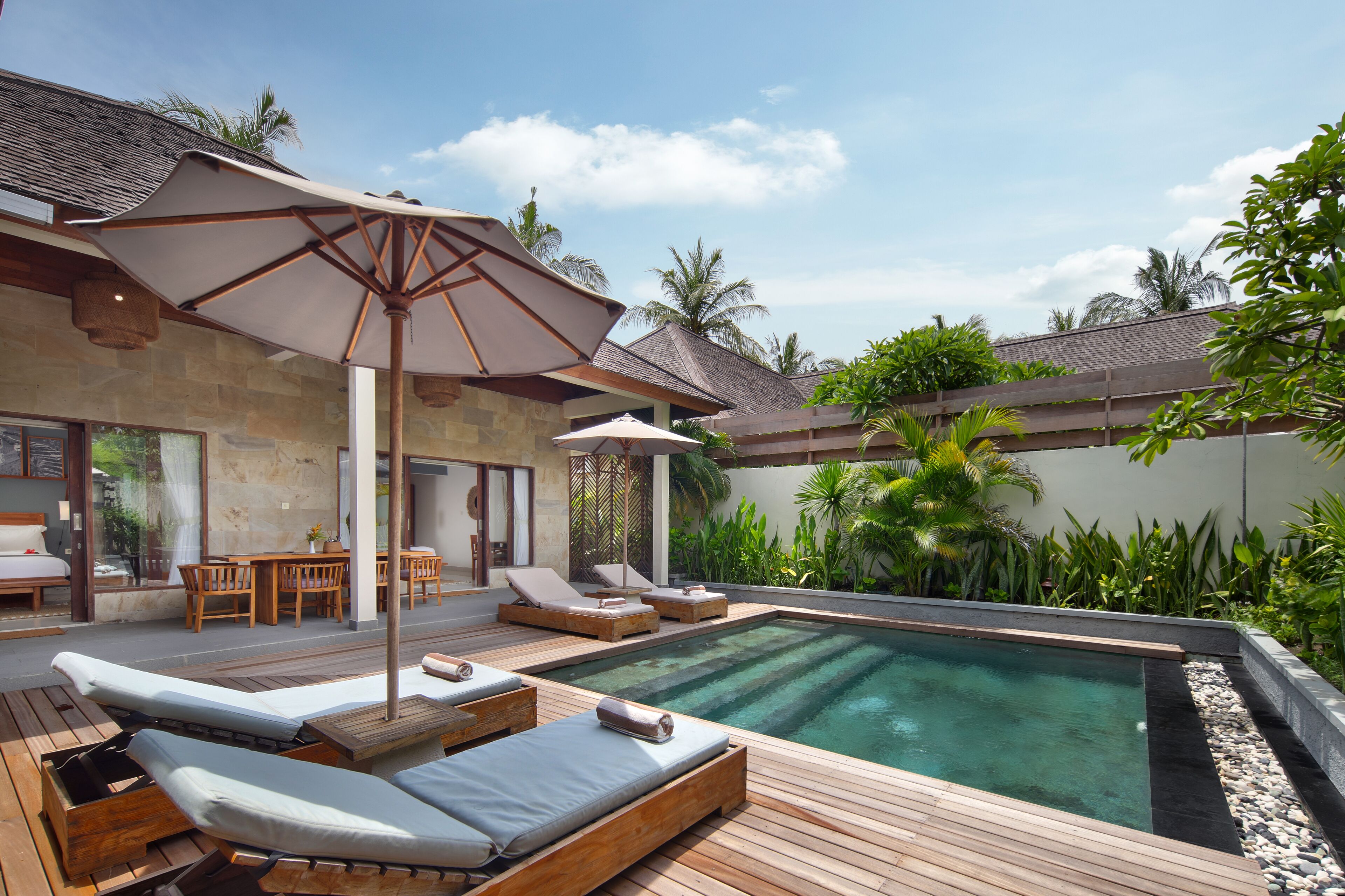 A poolside area with lounge chairs and an umbrella, surrounded by lush greenery and a stone building.