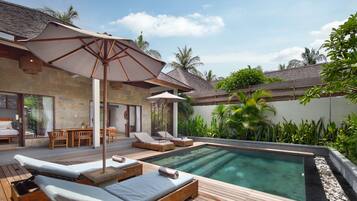 A poolside area with lounge chairs and an umbrella, surrounded by lush greenery and a stone building.