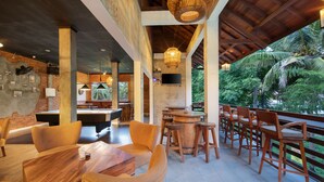 A spacious indoor area with a pool table, wooden tables, and chairs. There are large windows offering a view of lush greenery outside. - Utara Villas (Gili Trawangan)