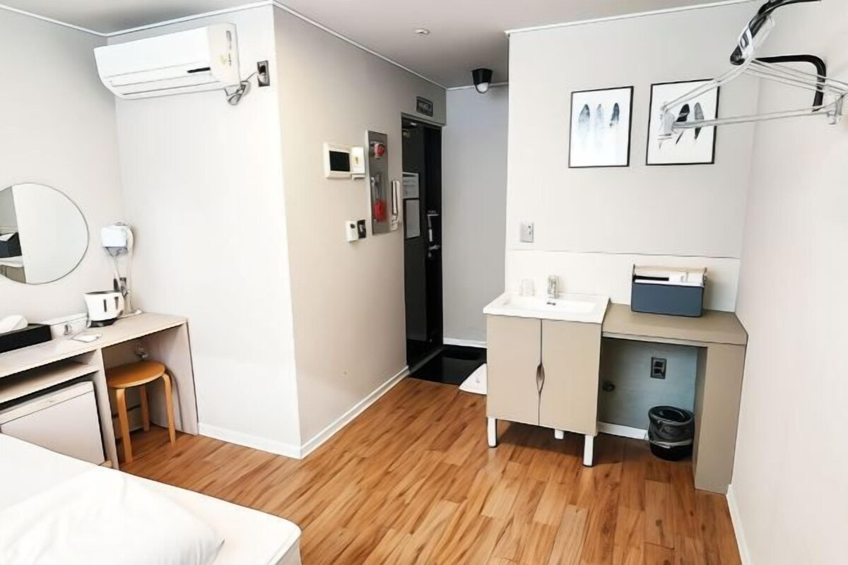 A modern hotel room with a wooden floor, a bed, a desk with a chair, a small stool, a vanity with a mirror, a sink, and a trash bin.