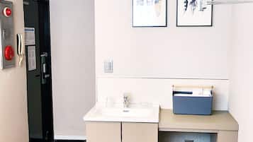 A compact hotel room with a kitchenette, a small desk with a sink, and a trash bin.