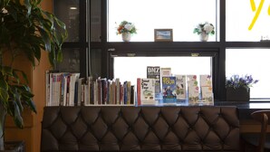 A seating area with a tufted leather couch, a bookshelf with travel books, and two flower vases on a window sill. - K- Grand Hostel Dongdaemun (Seoul)