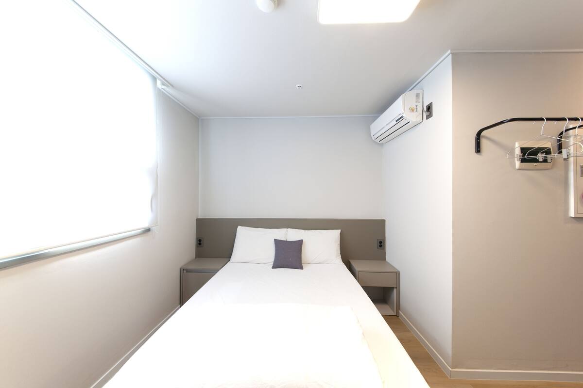 Una habitación de hotel con una cama, mesitas de noche, un equipo de aire acondicionado y una ventana.