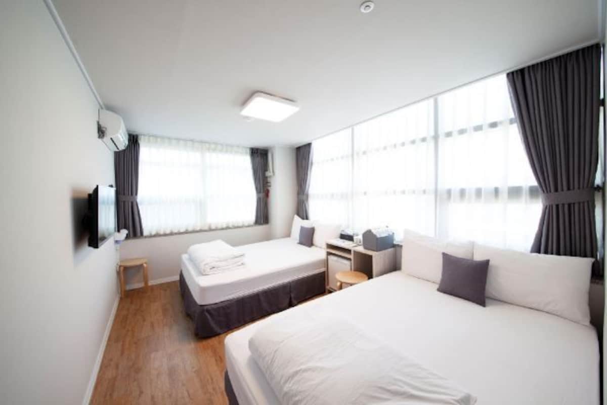A hotel room with two beds, a desk, a chair, a flat-screen TV, and large windows with curtains.