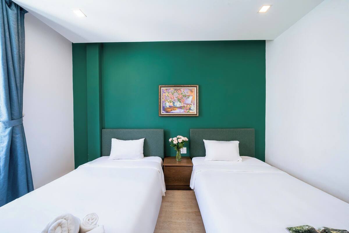 Une chambre d’hôtel avec deux lits, un mur d’accent vert, un tableau et une table de chevet agrémentée d’un vase de fleurs.