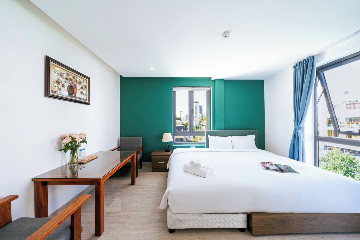 Une chambre d’hôtel avec un grand lit, un bureau avec une chaise, une petite table avec un vase de fleurs, une table de chevet avec une lampe et une fenêtre donnant sur la ville.