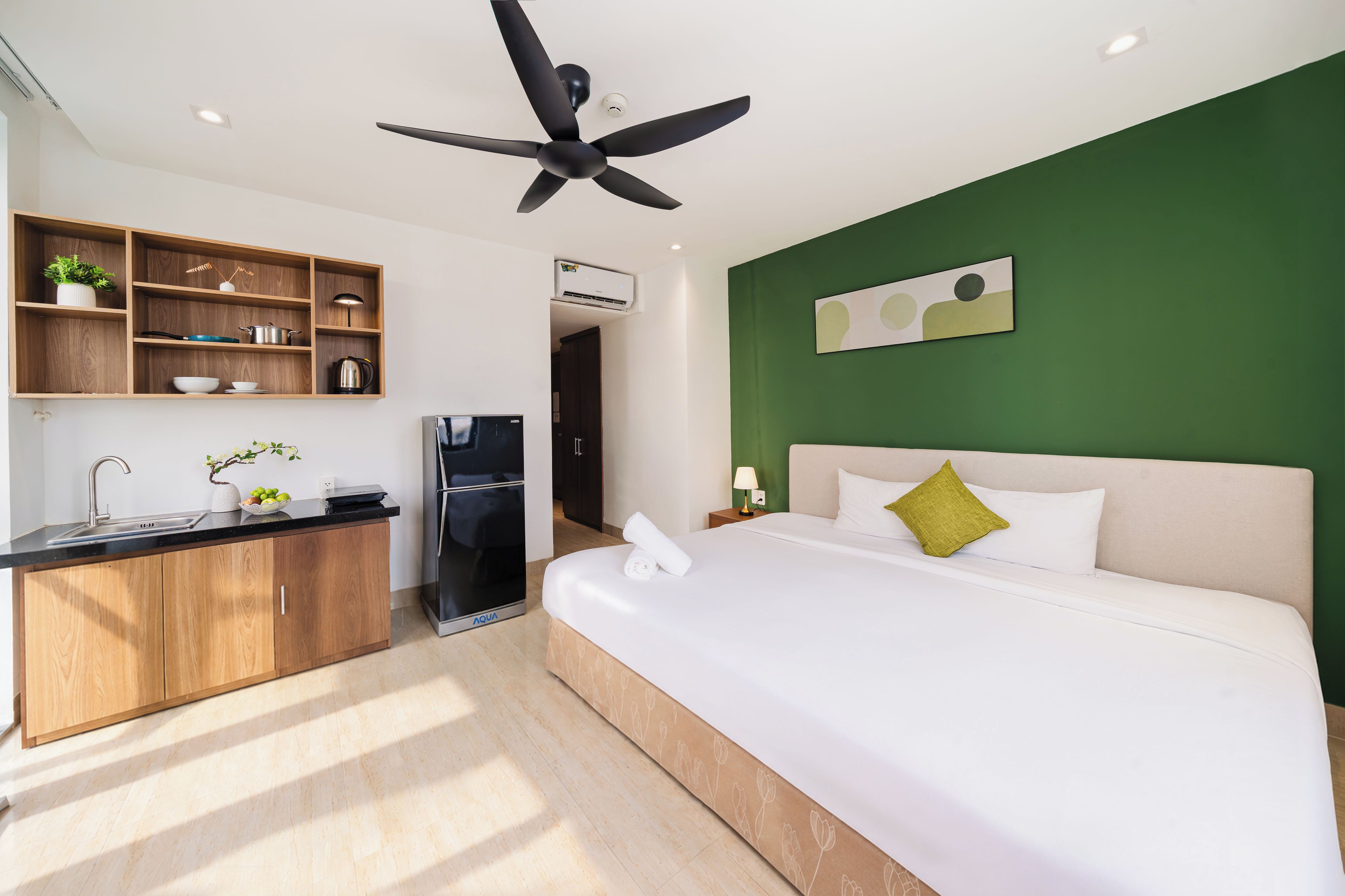 A hotel room with a bed, a kitchenette, a ceiling fan, and a green accent wall.