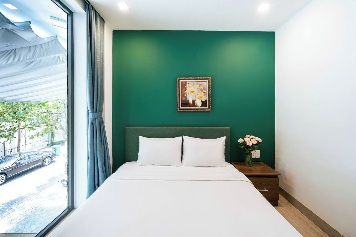 Une chambre d’hôtel avec un grand lit, un mur vert, un tableau encadré et une table de chevet ornée d’un vase de fleurs.