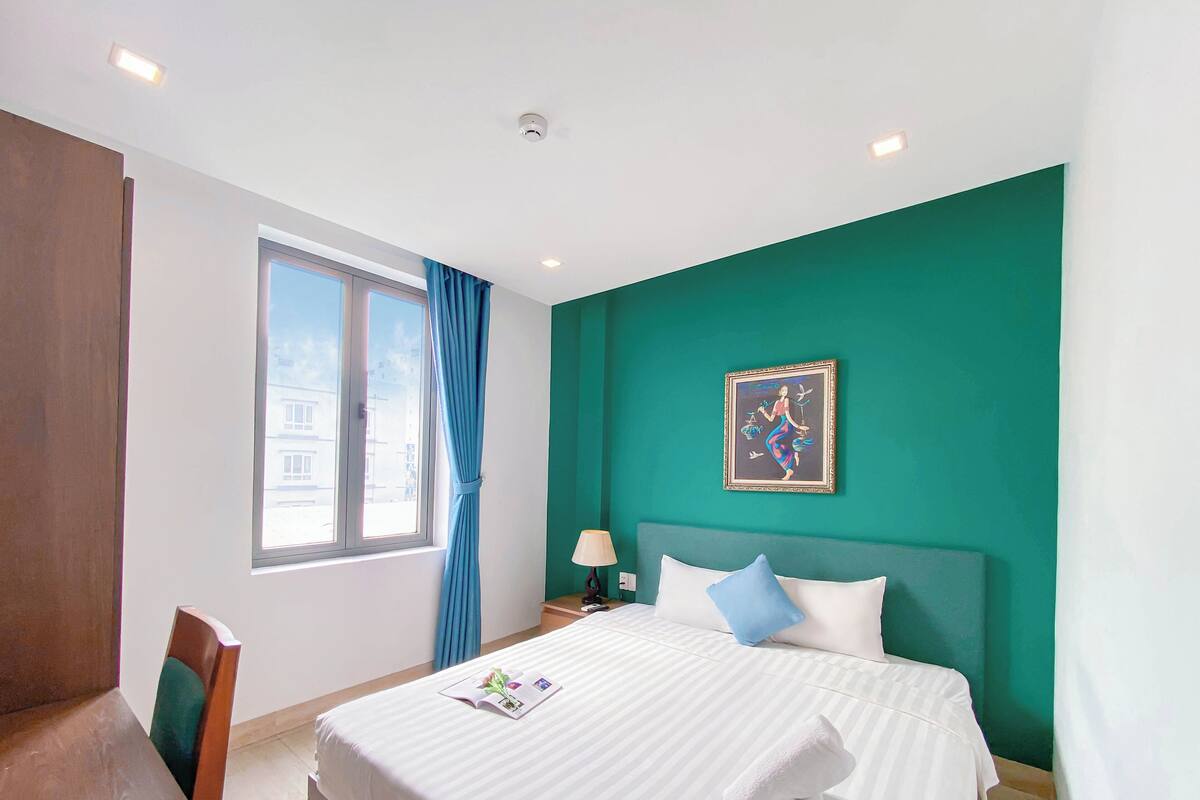 Une chambre d’hôtel avec un grand lit, une fenêtre avec des rideaux bleus, un tableau au mur et une chaise en bois.