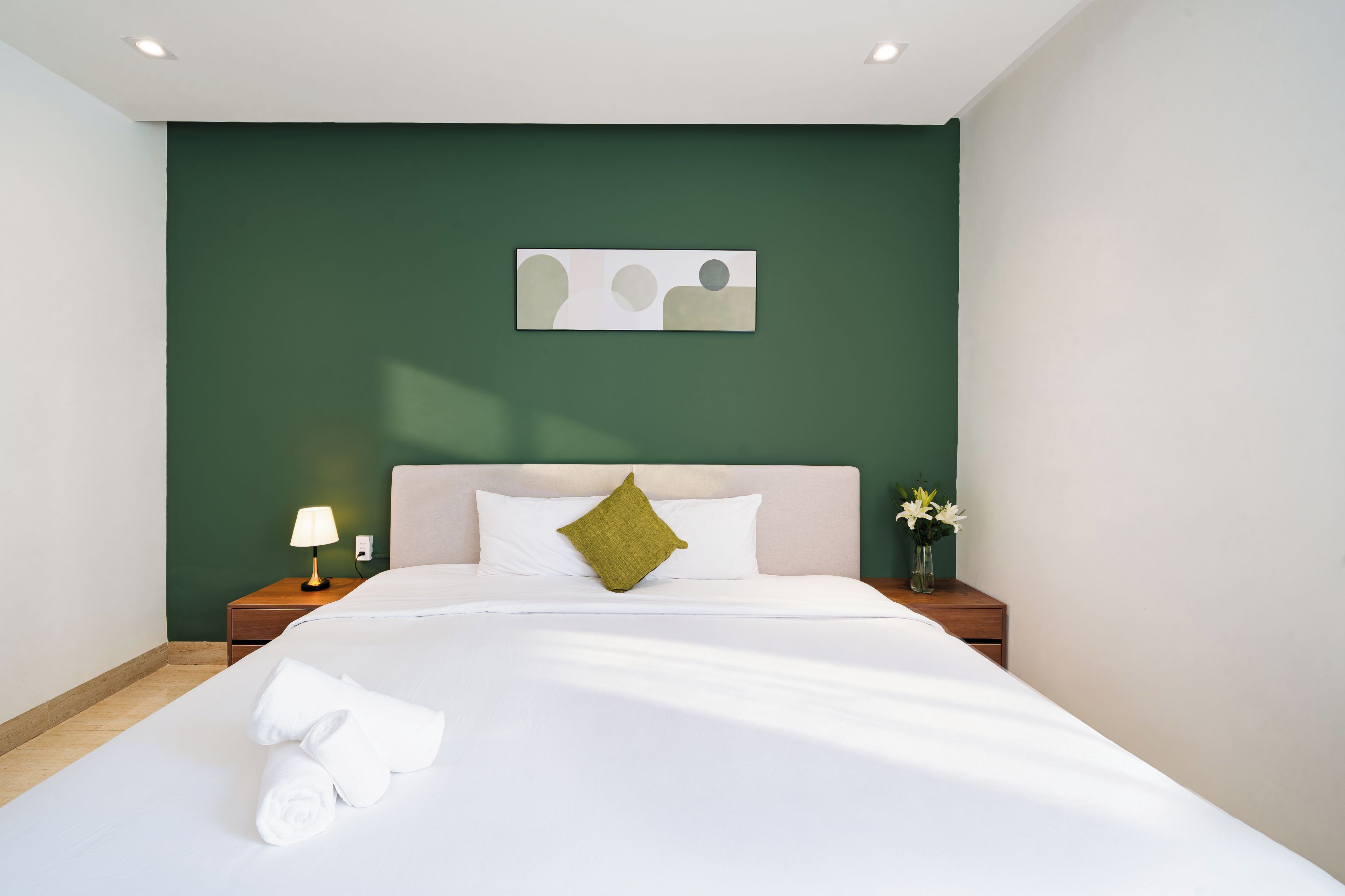 A neatly made bed with white linens and a green pillow, a bedside table with a lamp, and a wall-mounted artwork.