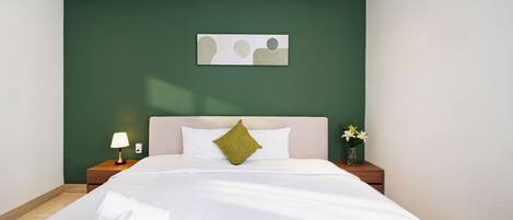 A neatly made bed with white linens and a green pillow, a bedside table with a lamp, and a wall-mounted artwork.