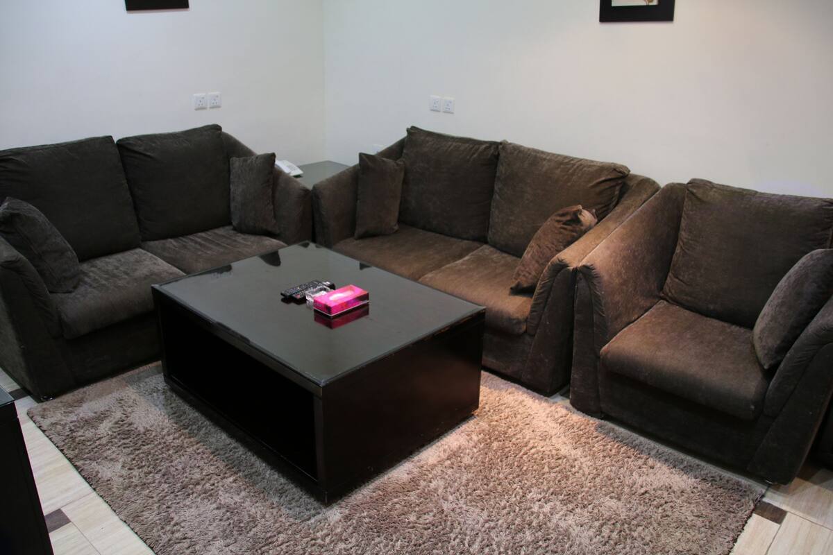 Ein Wohnzimmer mit einer dunkelbraunen Sofagarnitur, einem schwarzen Couchtisch und einem hellbraunen Teppich.