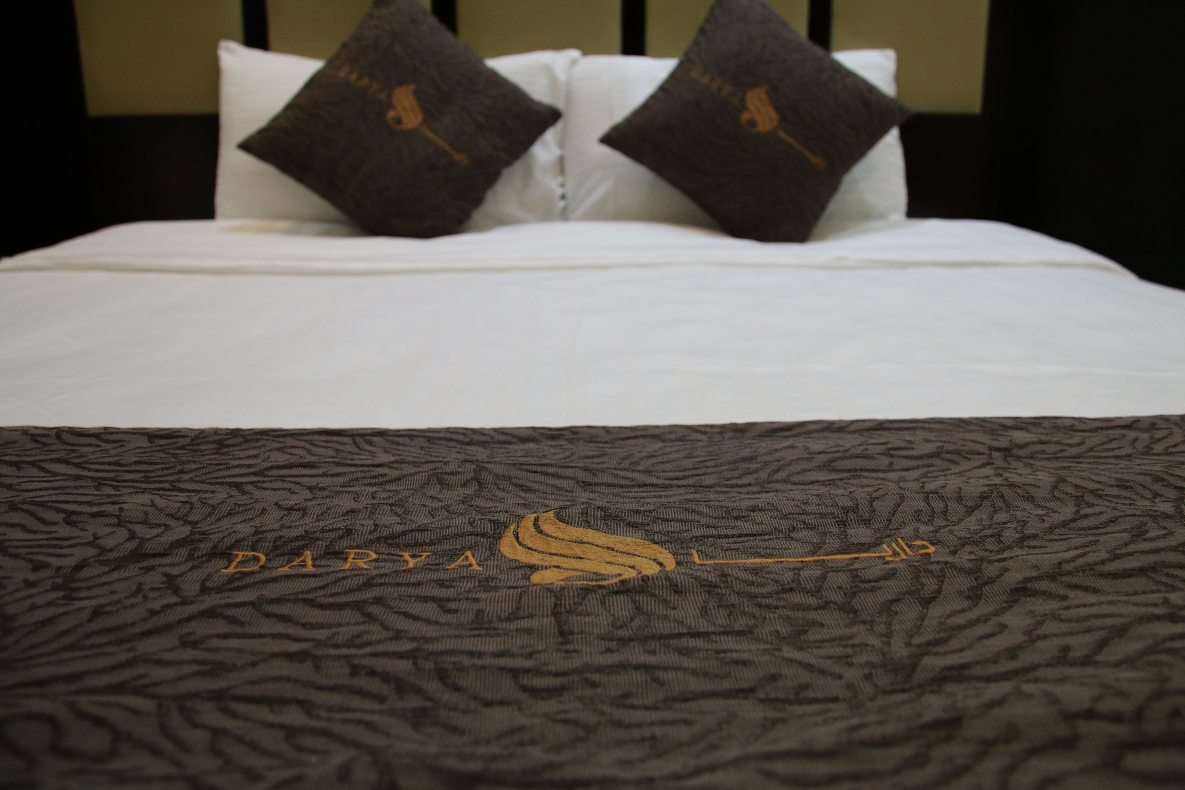 A bed with white sheets and two dark brown pillows. The bedspread has a textured pattern with the text "DARYA" and a logo.