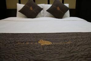 Una cama con sábanas blancas y dos almohadas marrón oscuro. El edredón tiene un diseño texturizado con el texto “DARYA” y un logotipo.