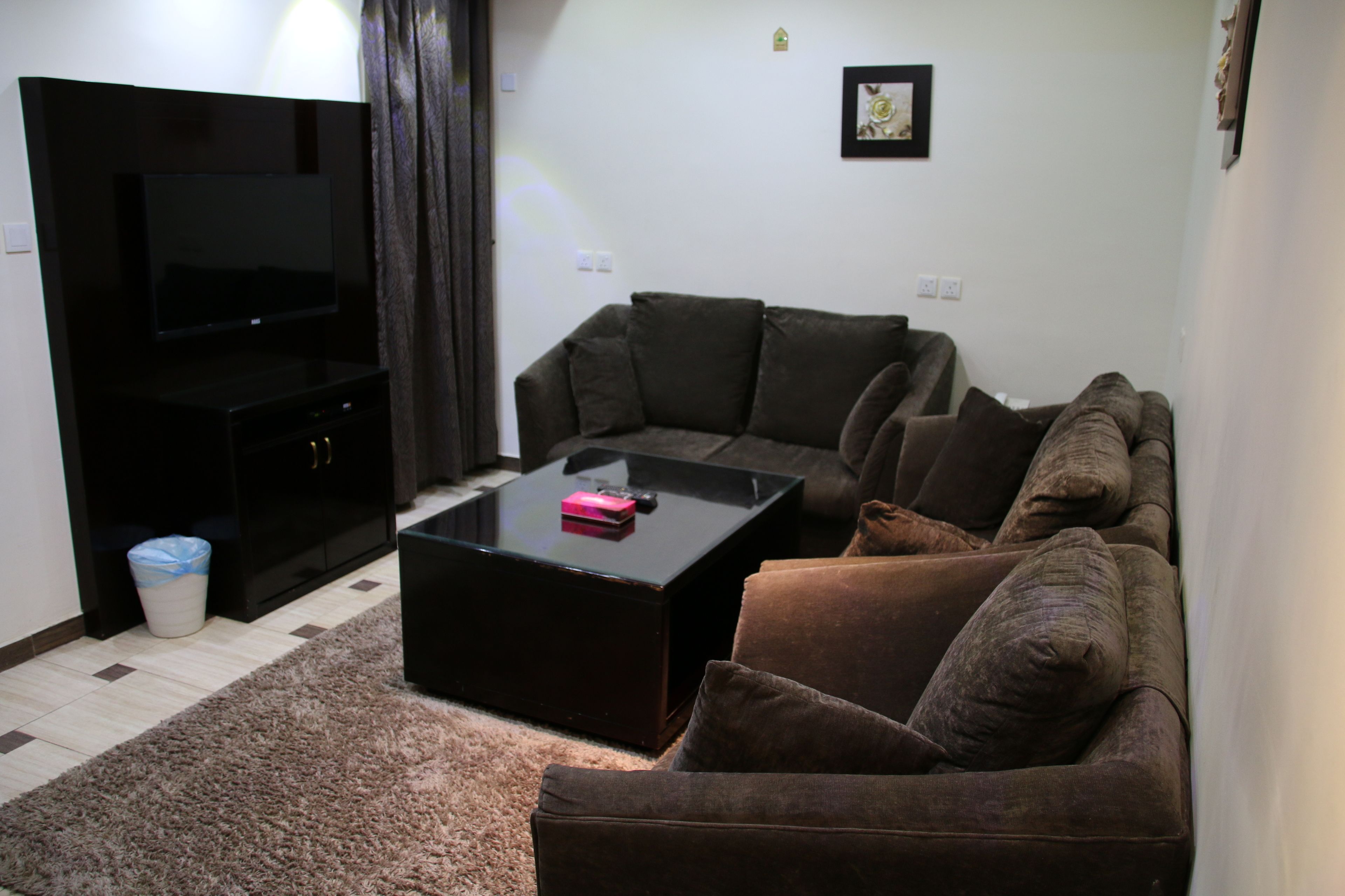 A living room with a sectional sofa, a flat-screen TV, a coffee table, and a trash bin.