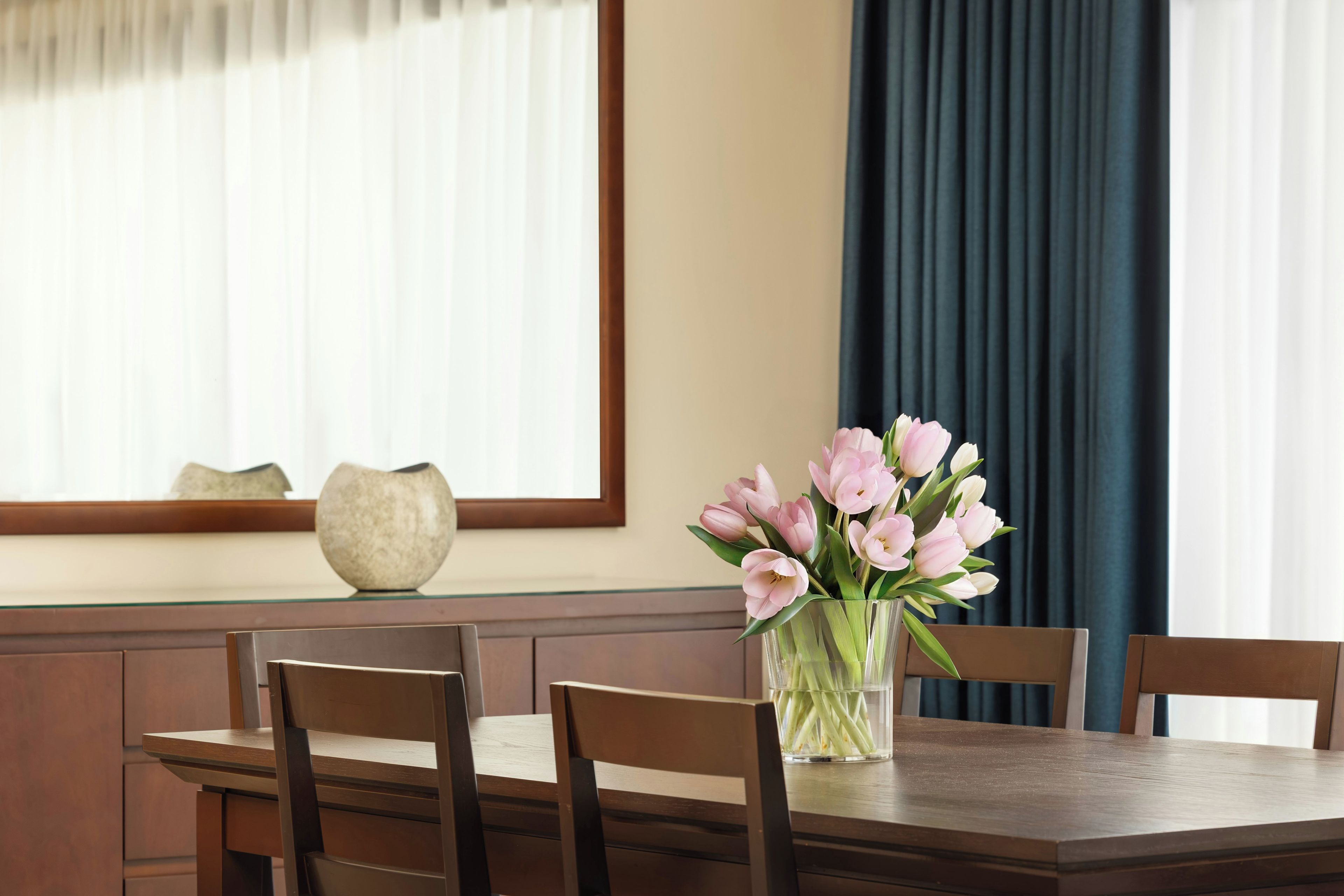 A dining table with a vase of flowers, wooden chairs, and a window with curtains.