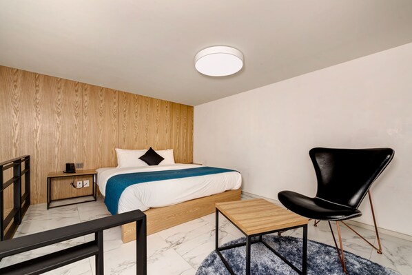 A modern bedroom with a wooden headboard, a bed with white bedding and a blue throw, a black leather chair, a wooden nightstand, and a round ceiling light. - Jamsil Delight Hotel (Seoul)