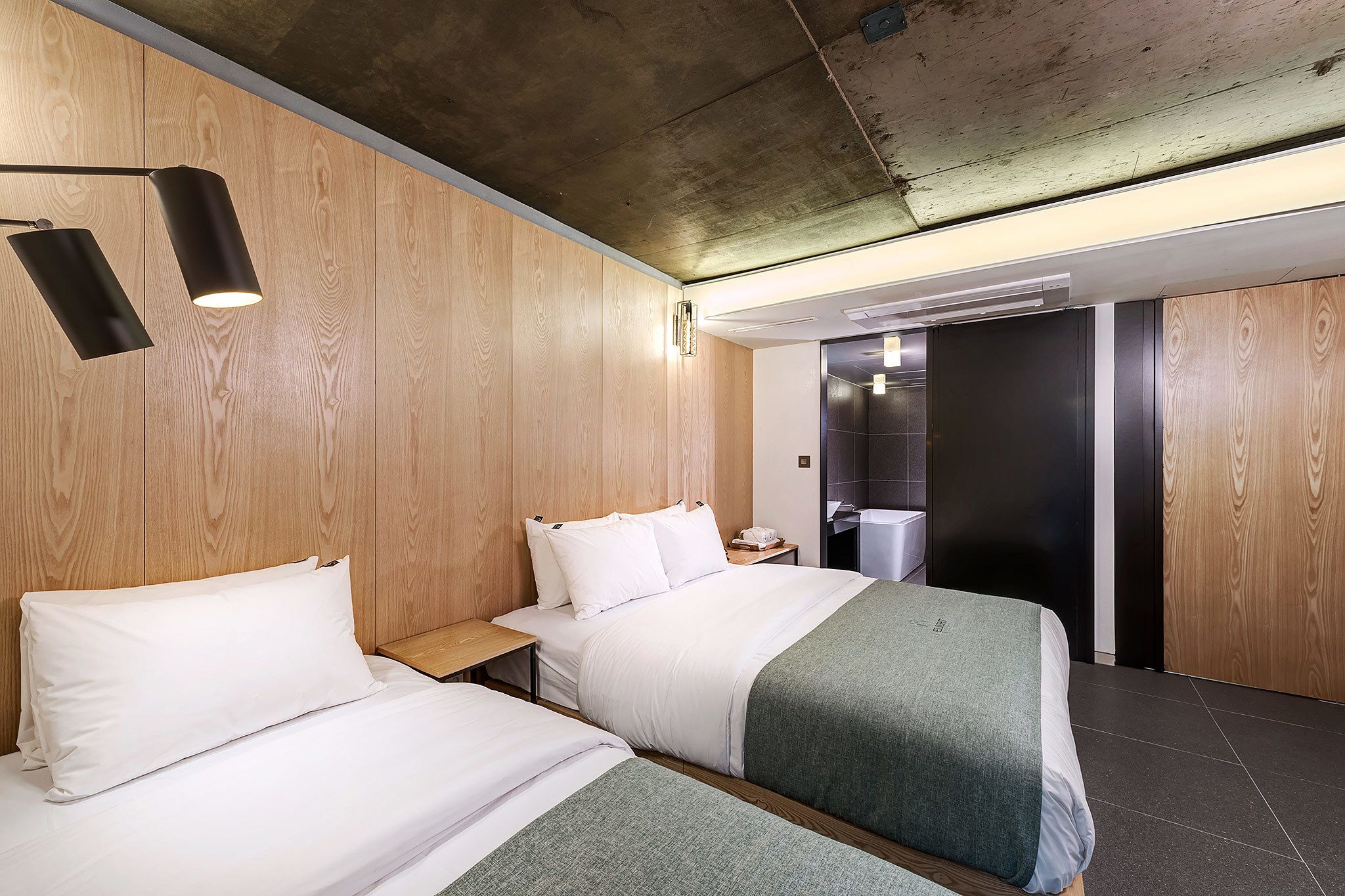 A hotel room with two beds, wooden panel walls, a concrete ceiling, and a modern design.