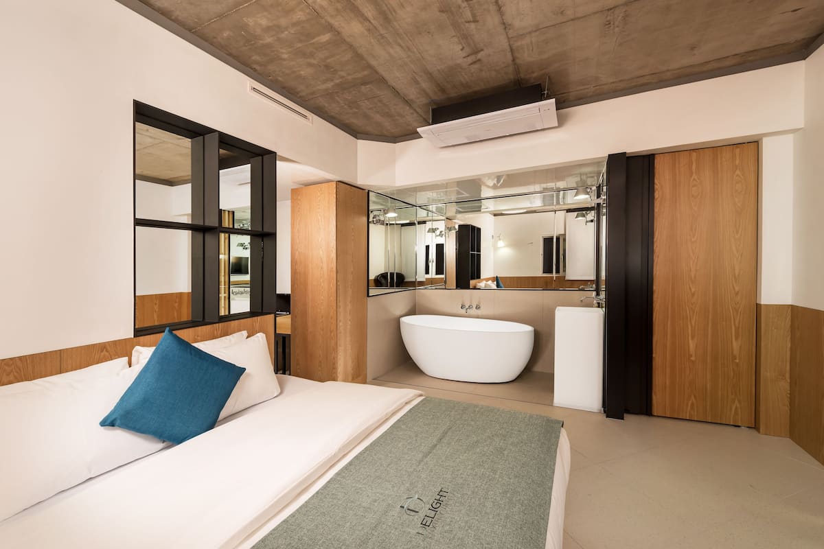 A modern bedroom with a large bed, a built-in window, a wooden headboard, a white bathtub, and a mirrored wall.