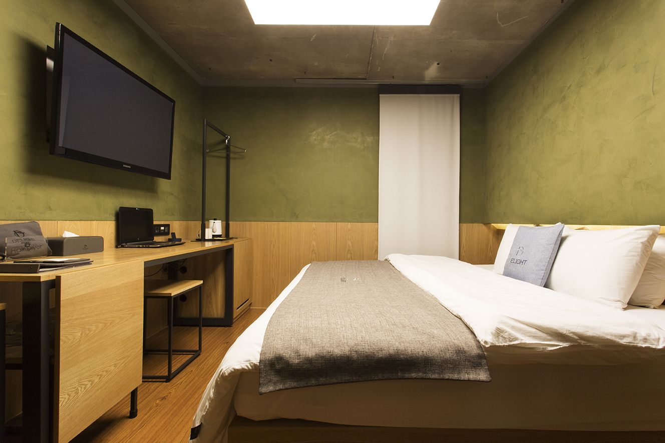 Hotellihuone, jossa on sänky, työpöytä tietokoneella, taulutelevisio ja ikkuna, jossa on verhot.