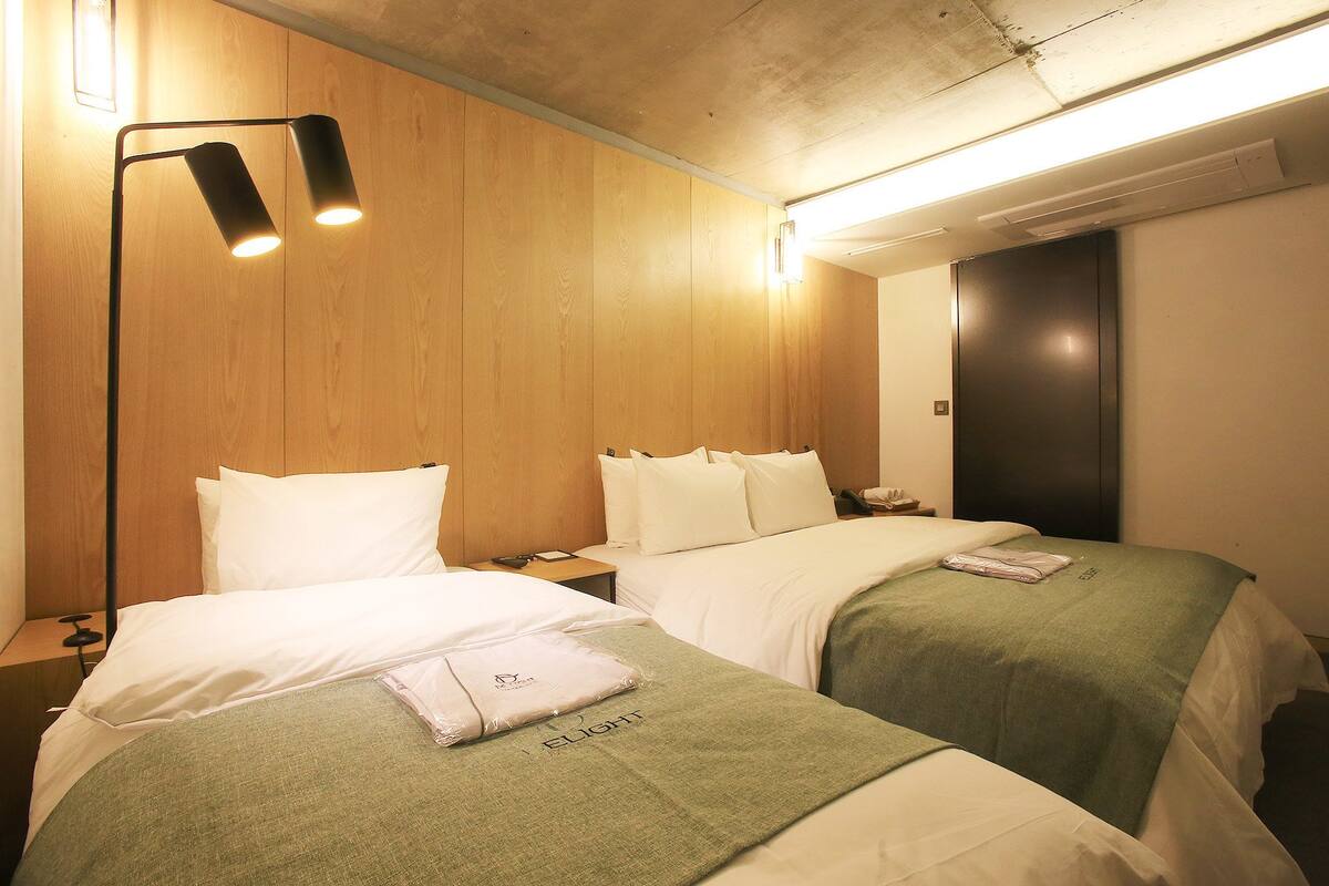 A hotel room with two beds, wooden paneling on the walls, a standing lamp, and a flat-screen TV.