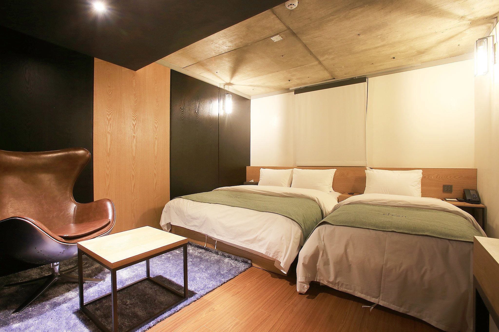 Habitación de hotel moderna con techo de madera, dos camas con ropa blanca, un sillón de cuero marrón, una mesita y una mesita de noche con un teléfono.