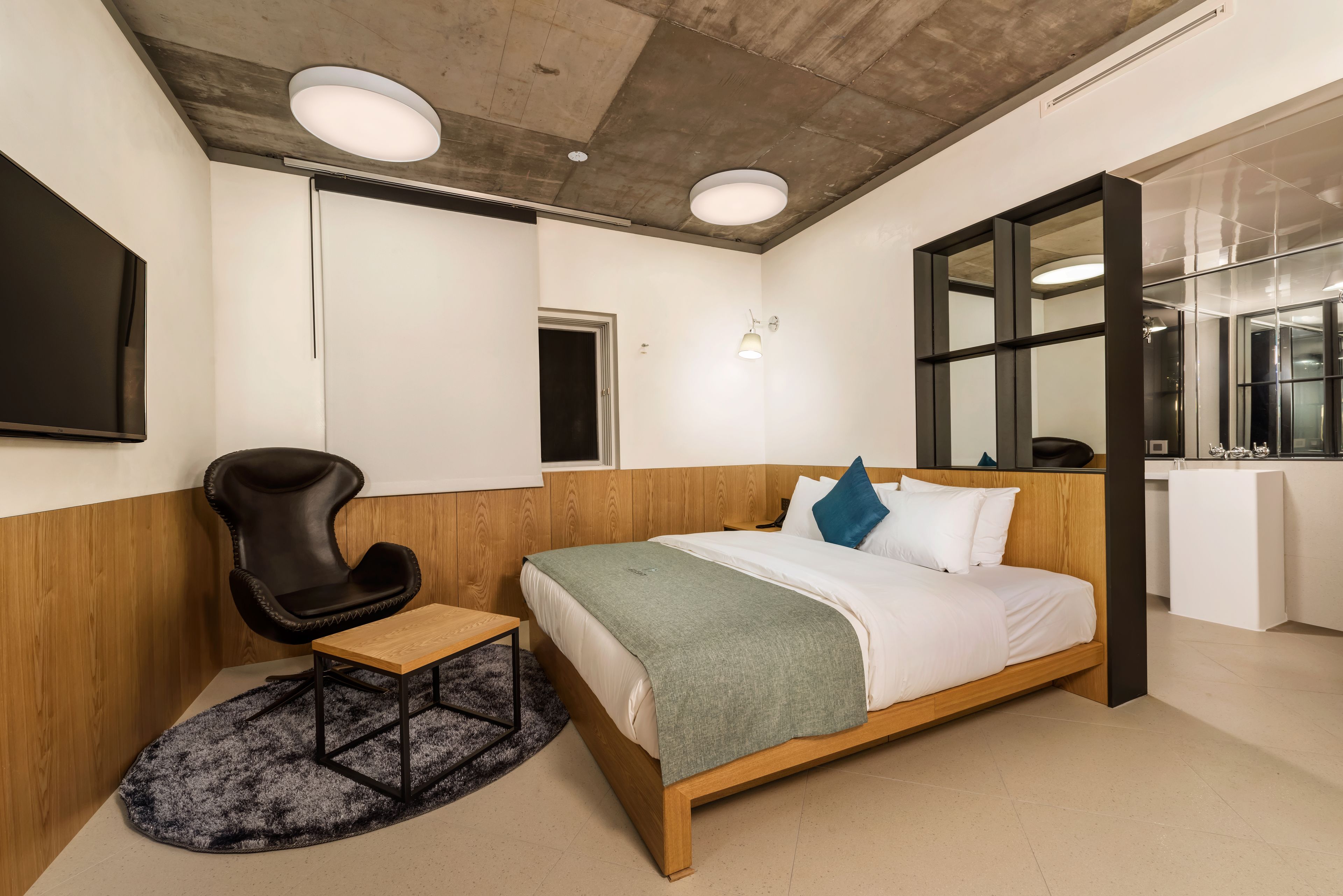 Una camera d'albergo moderna con un letto di legno, una poltrona in pelle nera, un tavolino e una grande finestra.