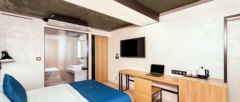 Habitación de hotel moderna con una cama, un escritorio, un televisor y un baño visible a través de una puerta abierta.