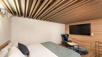 A modern bedroom with a wooden slatted ceiling, a bed with white bedding, a grey blanket, a black chair, and a flat-screen TV mounted on the wall.