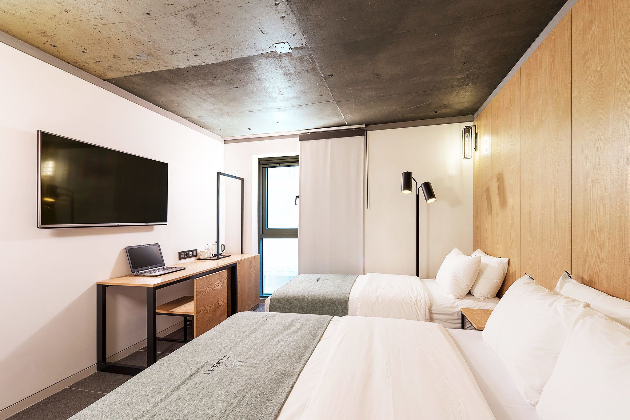 A modern hotel room with a large bed, a desk with a laptop, a flat-screen TV, and a wooden headboard.