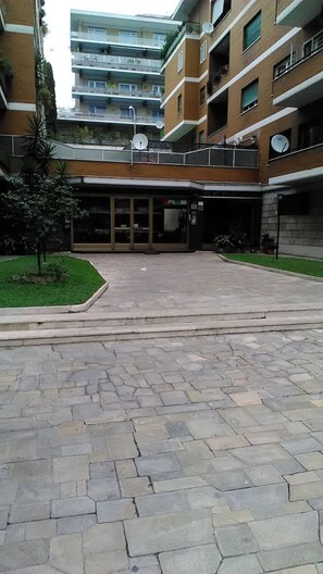 A courtyard with stone paving, a small tree, and a building entrance with glass doors. - Olimpica Relais (Rome)