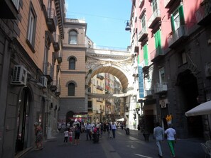 Une rue animée, bordée d’édifices historiques, dont une arche richement décorée et des immeubles à plusieurs étages dotés de balcons.