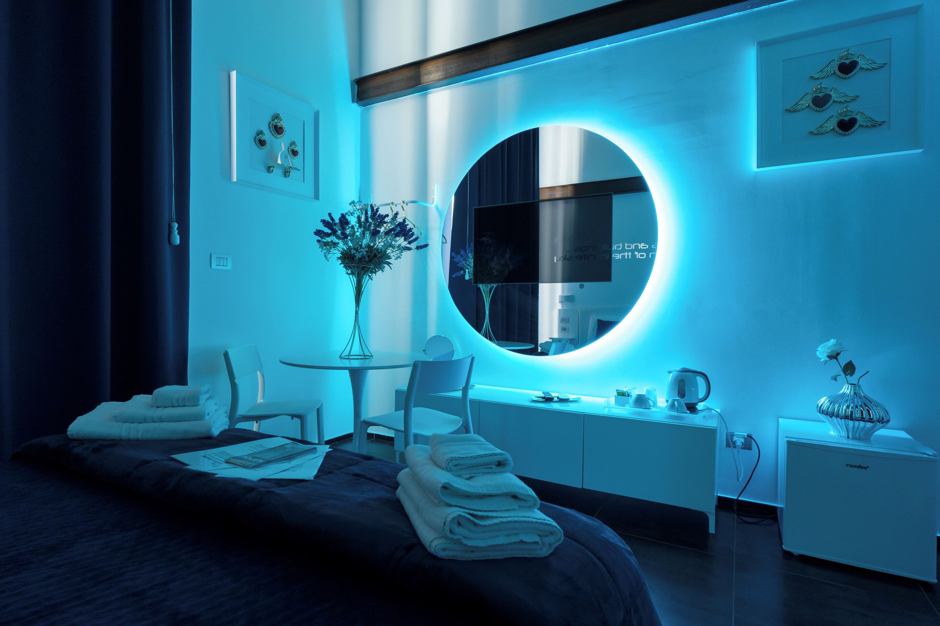 Quarto de hotel moderno com uma cama grande, um espelho redondo, uma escrivaninha com cadeira, uma mesinha com um vaso e uma mesa de cabeceira com abajur.