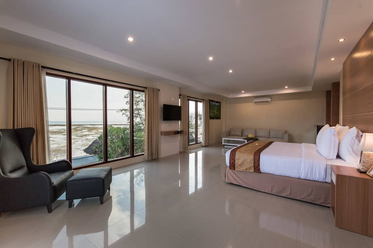 A modern hotel room with a large bed, a sofa, a TV, and a view of the beach.