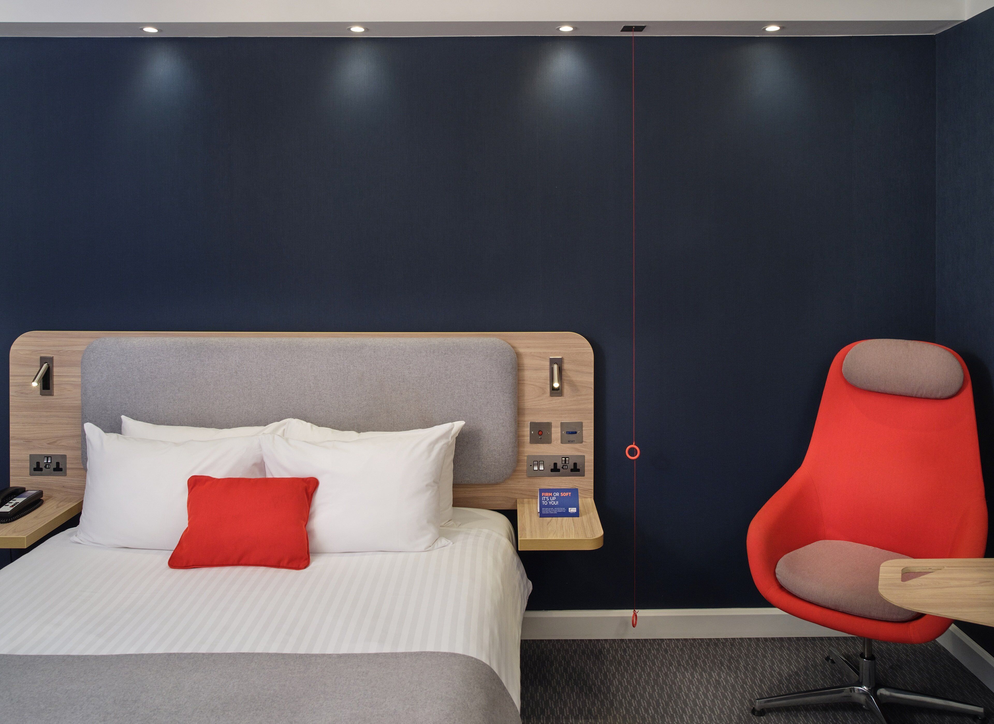 Une chambre d’hôtel avec un lit, une chaise rouge et un panneau de commande au mur.