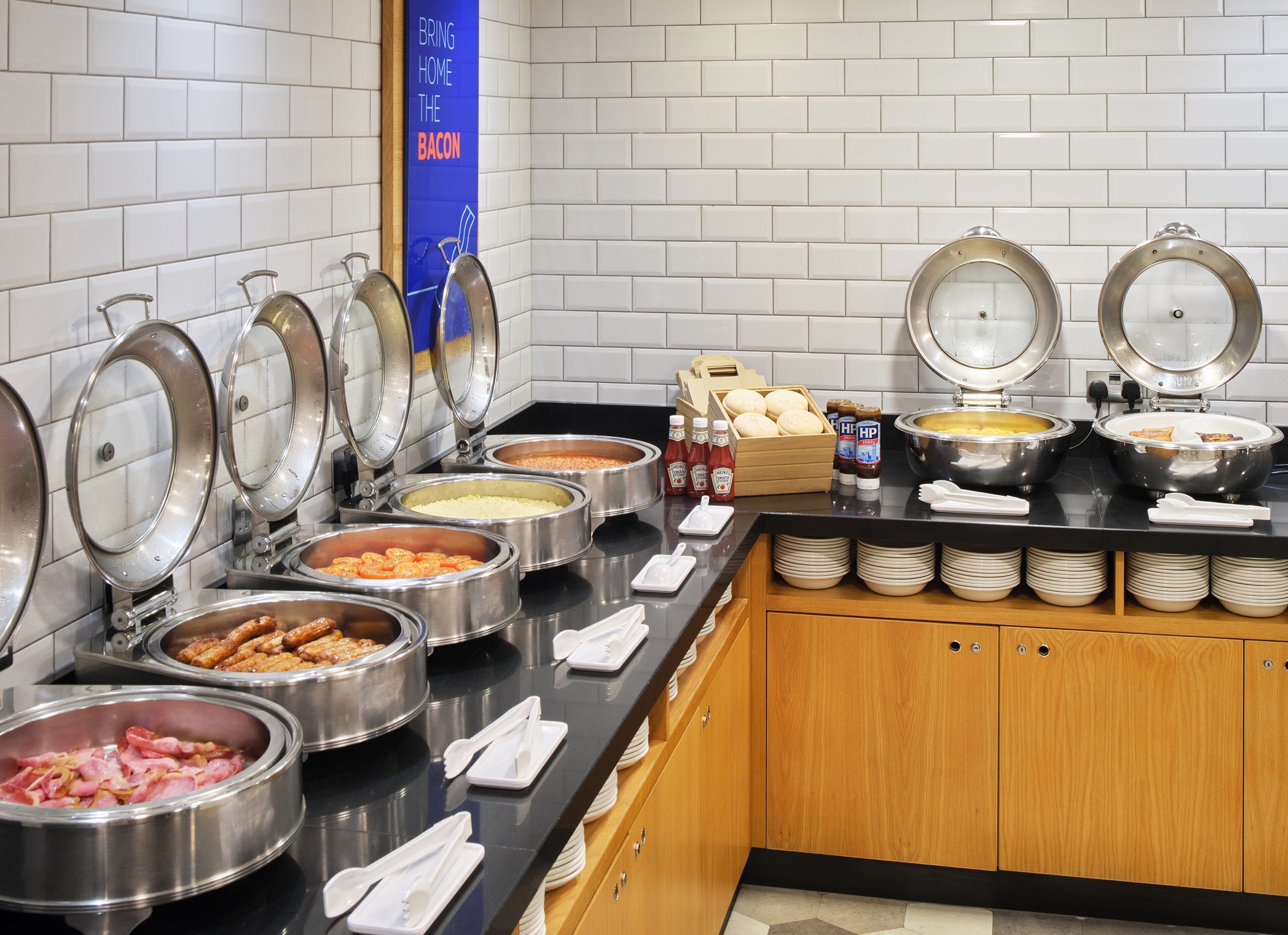 A buffet setup with various food items in metal containers, a condiment station, and a sign that reads "BRING HOME THE BACON".