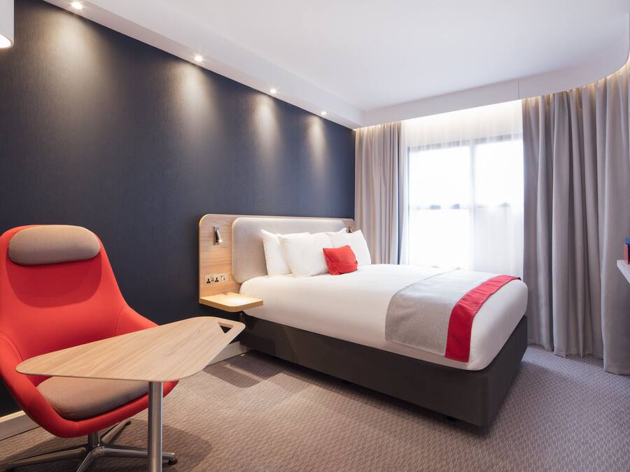Une chambre d’hôtel moderne avec un grand lit, un fauteuil rouge, une petite table et un téléviseur fixé au mur.