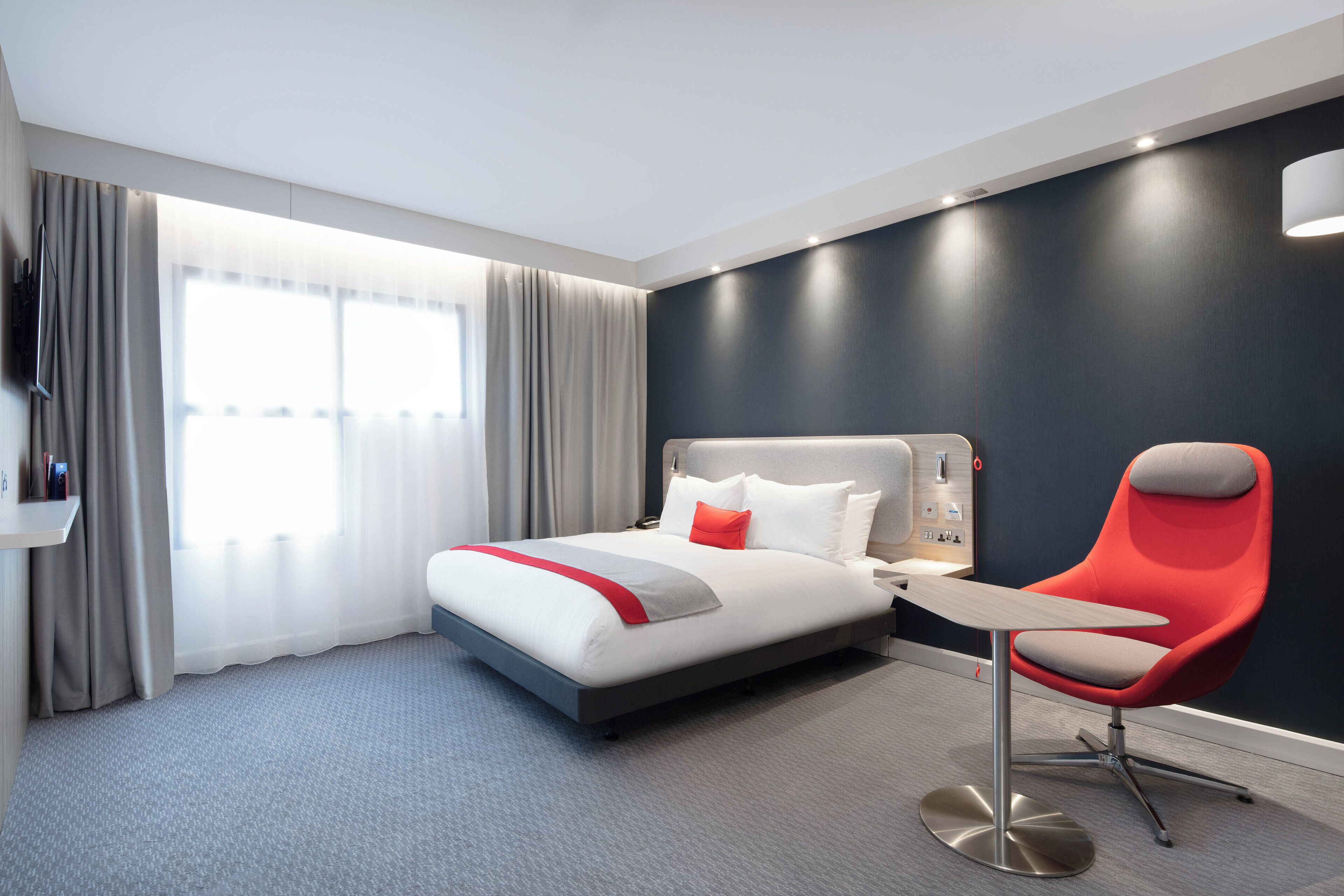 Una habitación de hotel moderna con una cama, una silla roja, una mesita y un televisor.