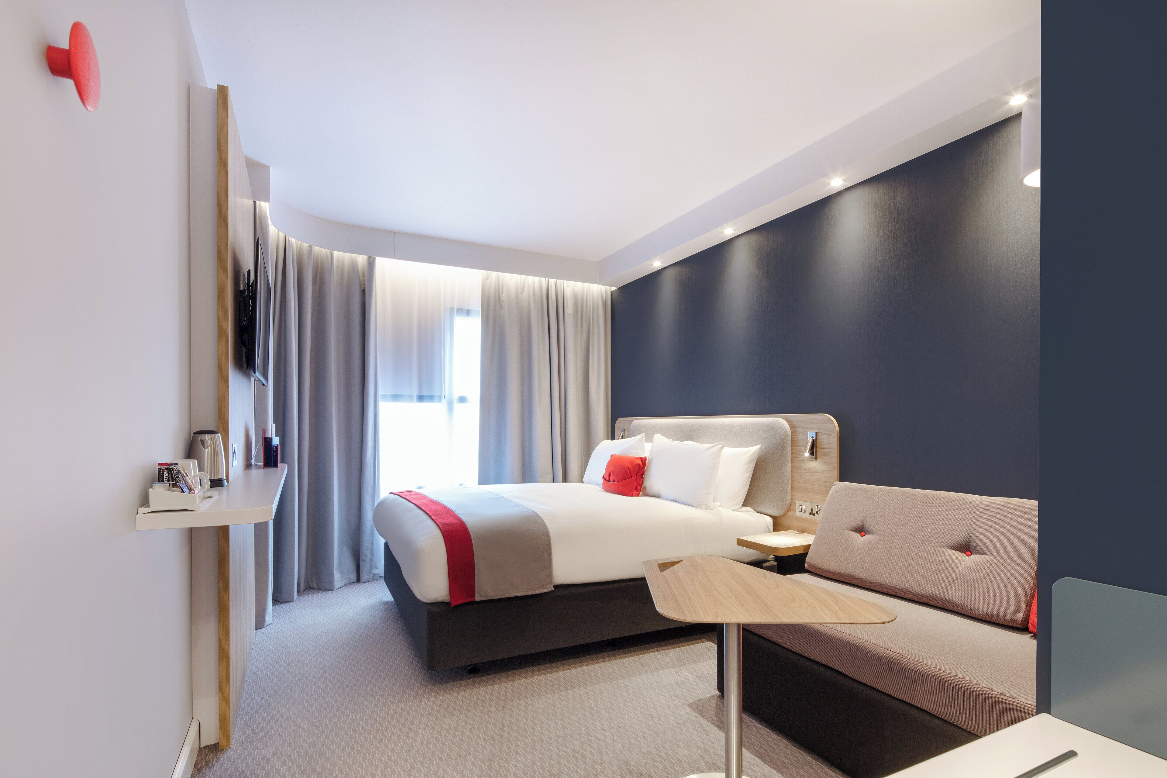 A modern hotel room with a bed, a sofa, a small table, a TV, and a window with curtains.