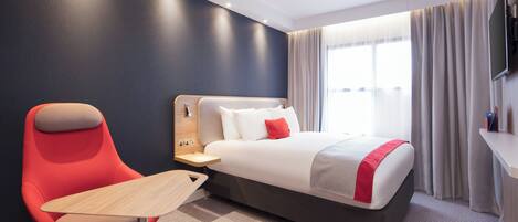 A hotel room with a bed, a red chair, a small table, a TV, and a window with curtains.