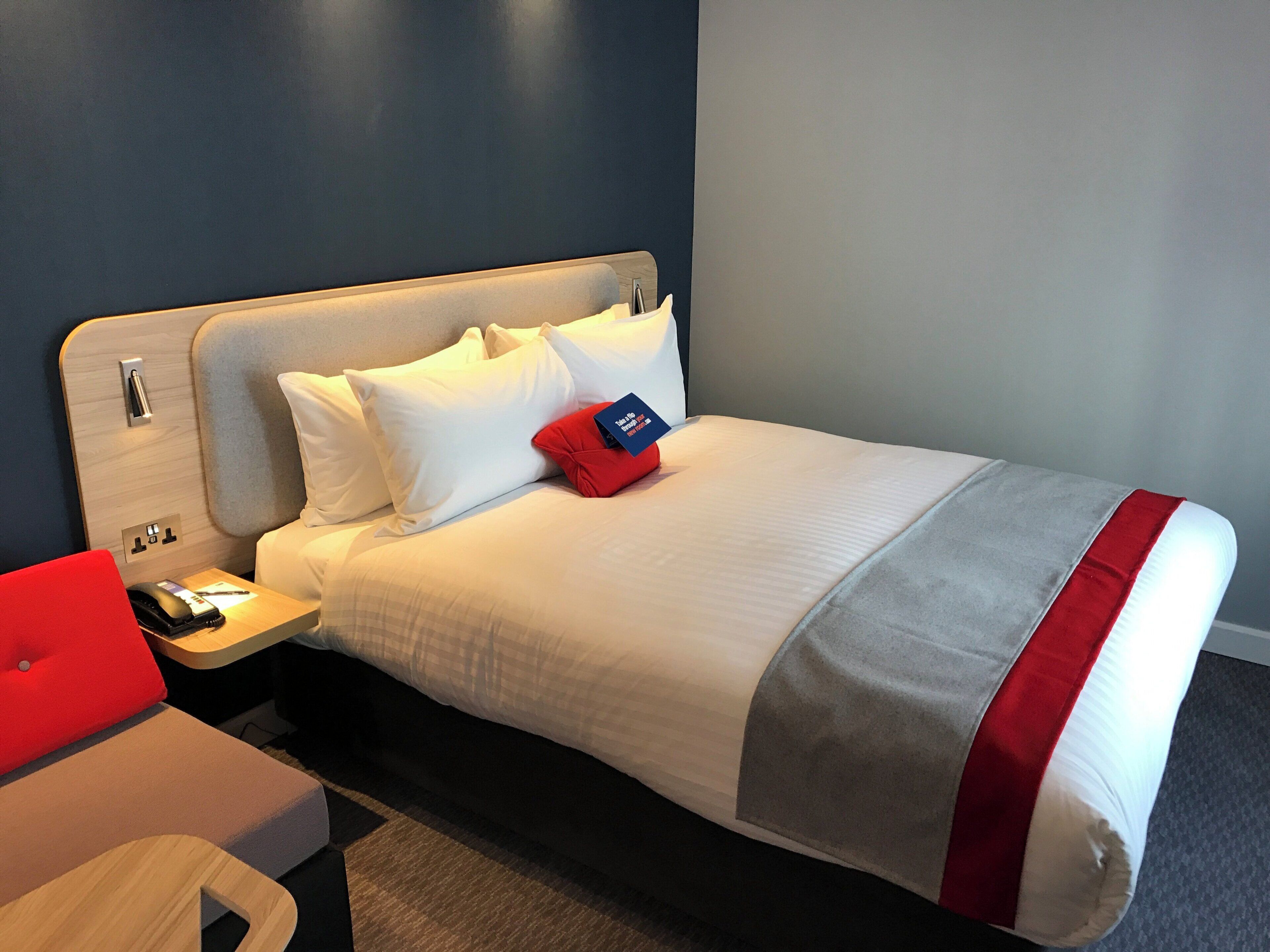 A hotel room with a bed, a nightstand, a red chair, and a wooden side table.
