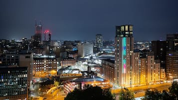 Un paisaje urbano nocturno con edificios iluminados, un rascacielos destacado con espectáculos de luces verticales y una calle concurrida con vehículos.
