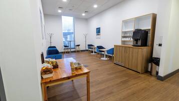 A modern waiting area with a wooden table, blue chairs, a coffee machine, and a view through a large window.