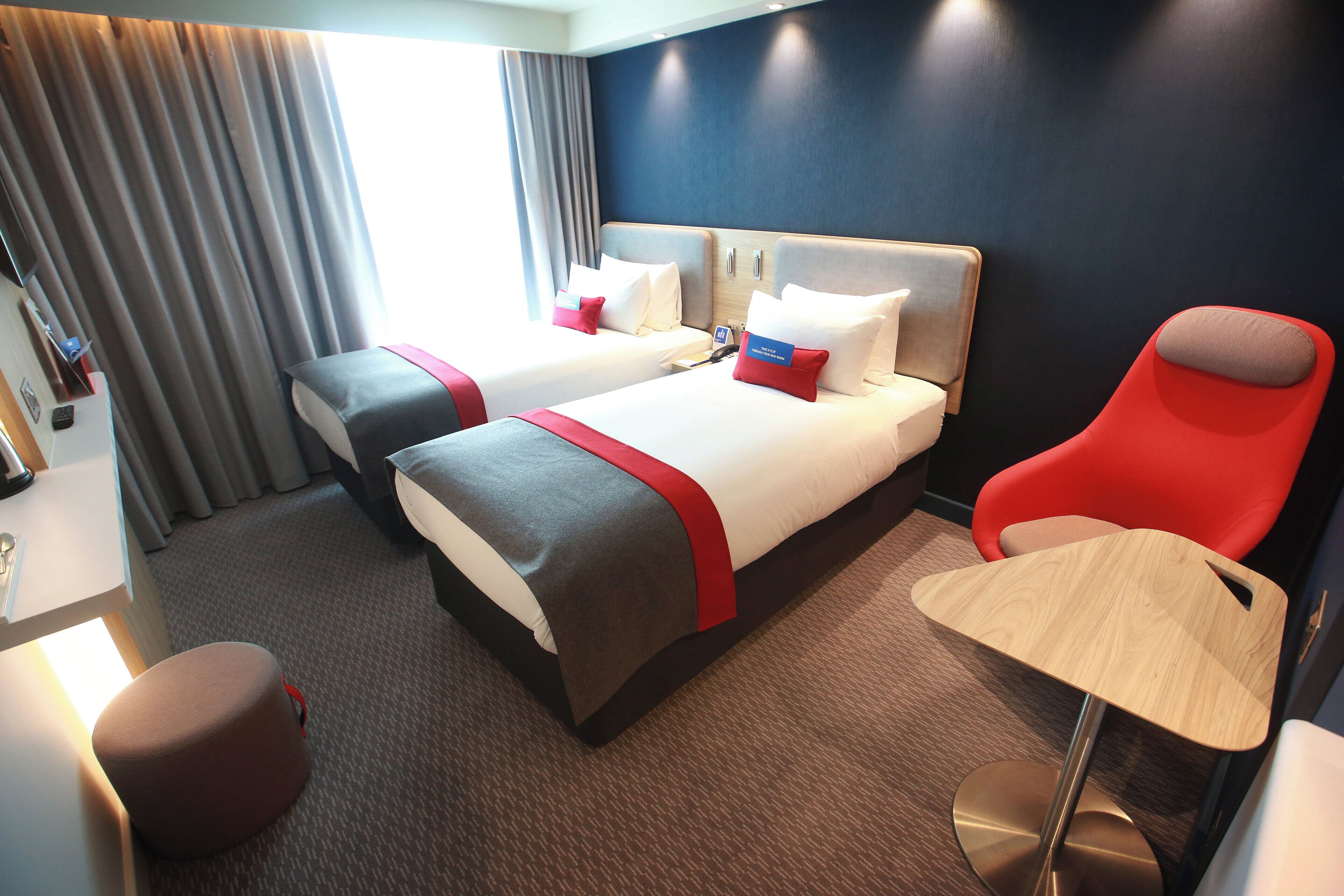 Habitación de hotel con dos camas, una silla roja, una mesita y un escritorio con lámpara.
