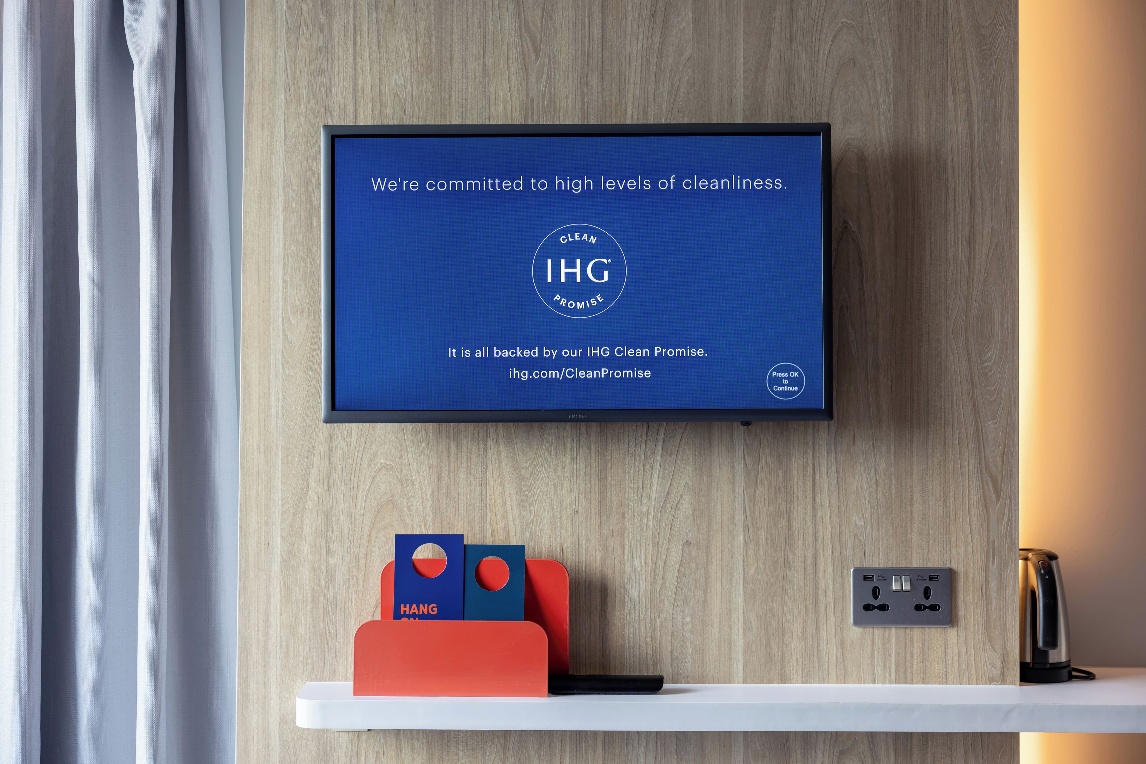 A TV displaying a message about cleanliness commitment, a wall-mounted power outlet, and a red and blue hanging organizer on a wooden wall.