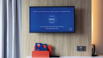 A TV displaying a message about cleanliness commitment, a wall-mounted power outlet, and a red and blue hanging organizer on a wooden wall.