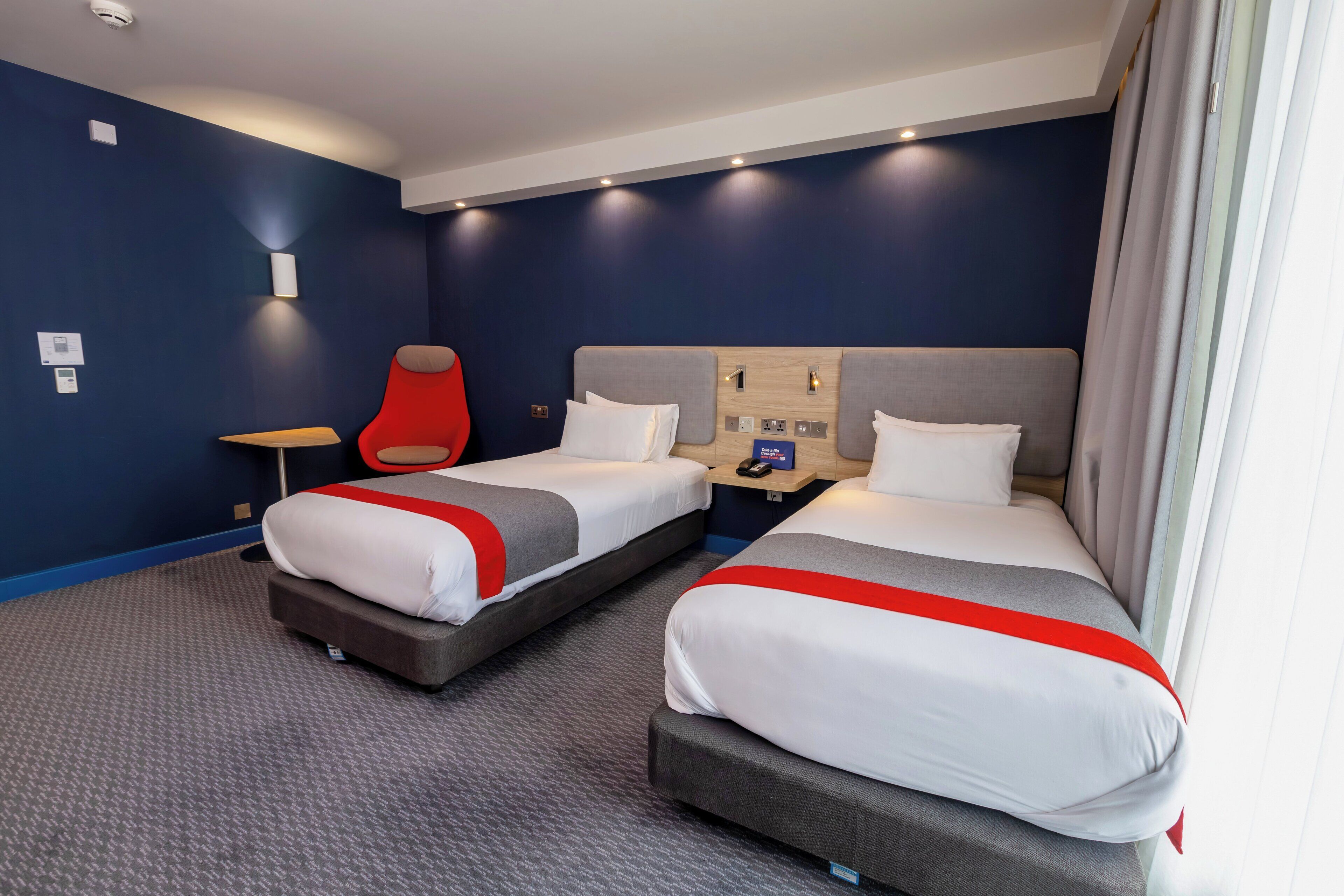 A hotel room with two beds, a red chair, a nightstand, and a wall-mounted control panel.