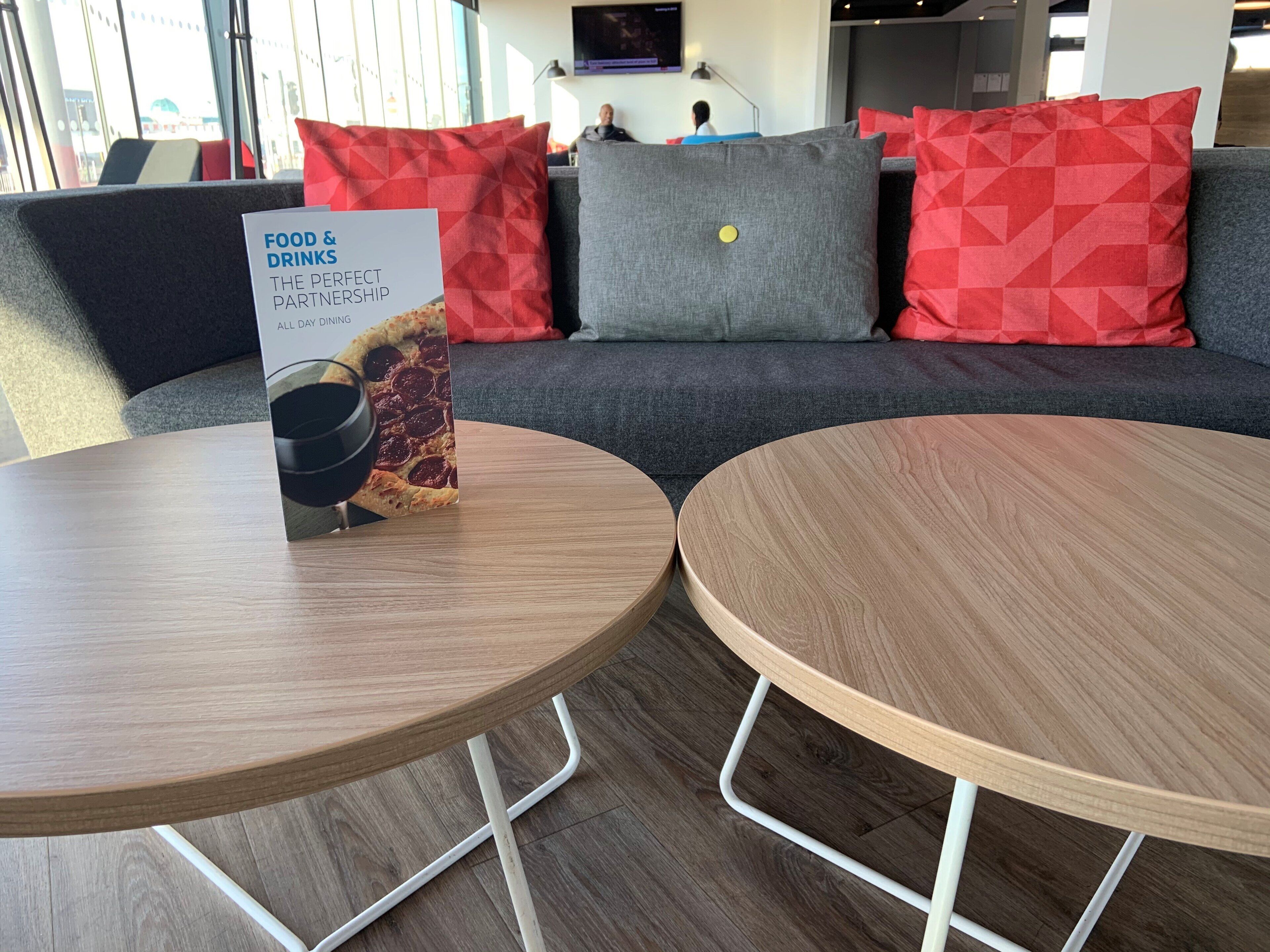 A modern interior with two round wooden tables, a sofa with red and grey cushions, and a sign for a food and drinks service.