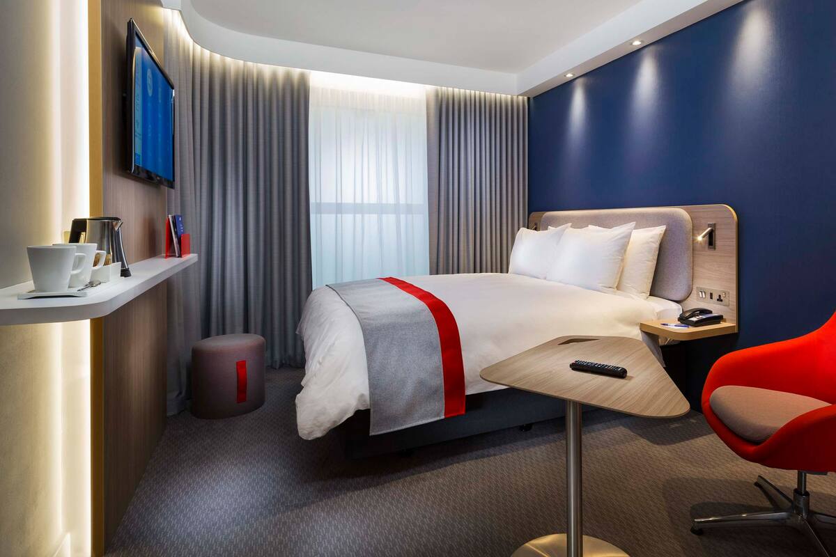Een moderne hotelkamer met een bed, een rode stoel, een bureau met een afstandsbediening, een kleine prullenbak en een plank met bekers en boeken.