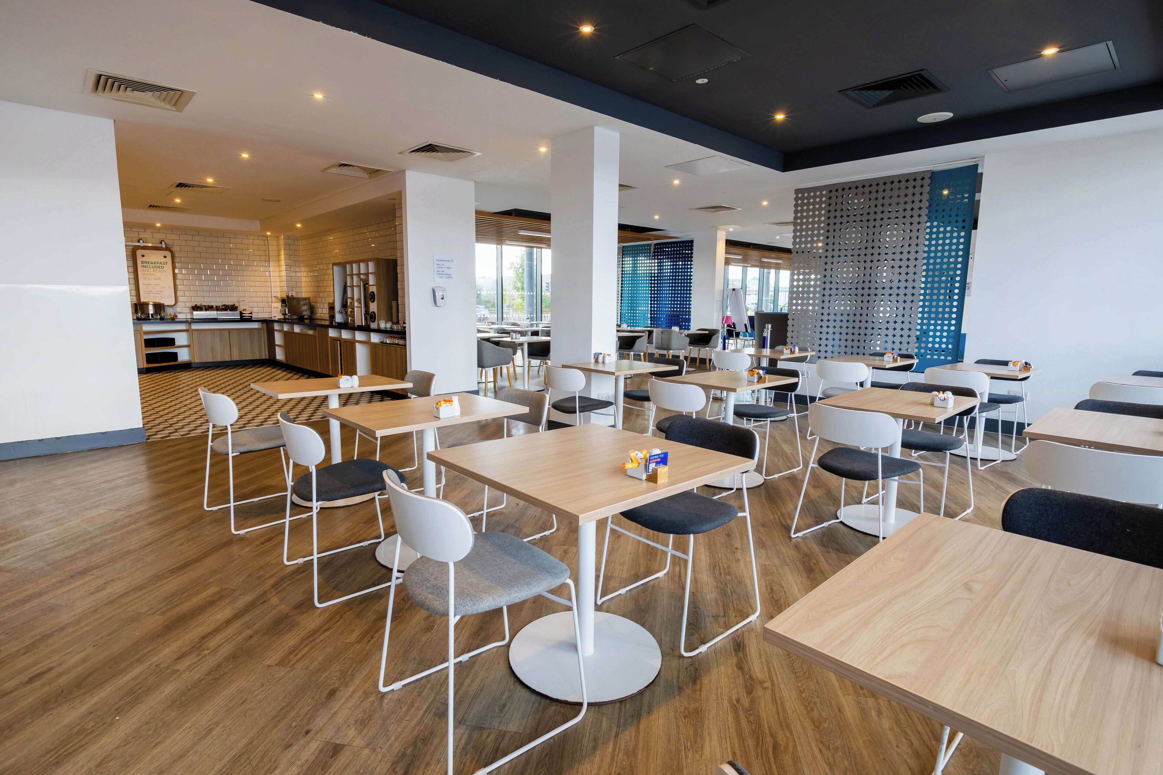 A modern dining area with wooden tables and chairs, a kitchen in the background, and a decorative wall.